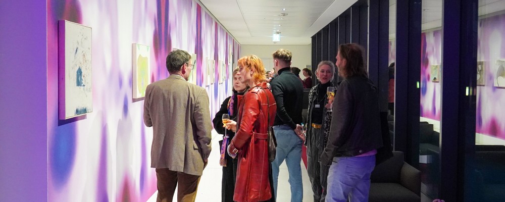 Mehrere Personen unterhalten sich und betrachten Gemälde, die an einer Wand mit einem Muster aus Rot-, Violett- und Lila-Tönen hängen. 